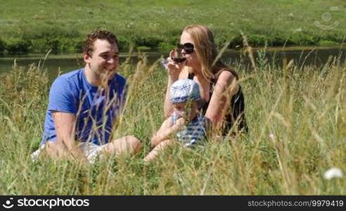 Mother, father, and little son sitting on the grass near the small river. Woman drinking wine, boy eating. Family picnic on a sunny summer day