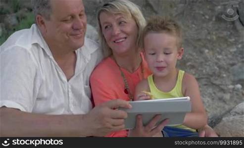 Mother, father, and son watching a video on a tablet while sitting on the beach. The boy is eating food, and the parents are talking.