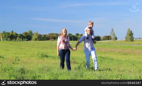 Mother, father, and their little son walking in the countryside. Couple holding hands, man kissing his wife while boy sitting on father's shoulders.