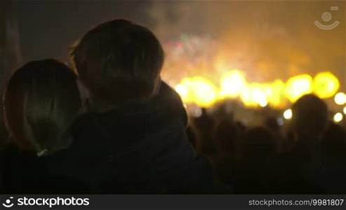 Mother holding son in arms and they enjoying bright firework show at night. Celebration in the city. Back shot