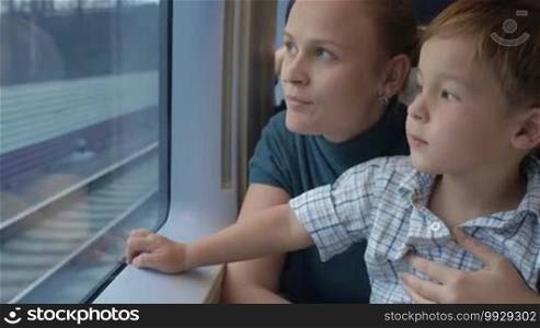 Mother holding son on the lap in moving train. She talking to the child while they looking out the window