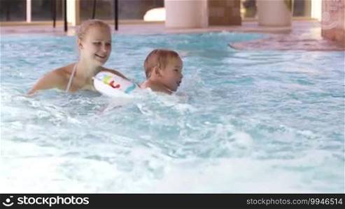 Mother laughing as she plays with her young son in a swimming pool as the child learns to swim using a plastic water ring