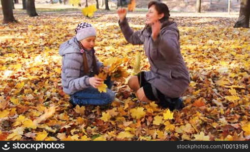 Mother with her daughter sit squatted and scatter yellow leaves in beautiful autumn city park