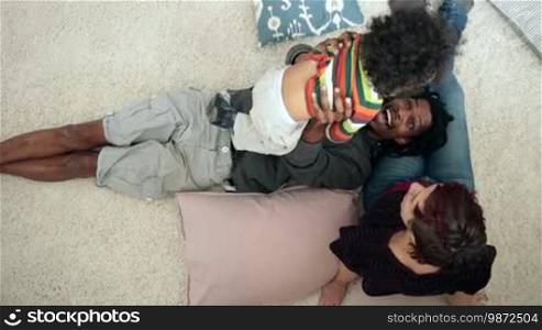Multiethnic family spending time and having fun together at home. Joyful African American father with dreadlocks raising his mixed-race toddler son up while lying on his back, his head on Caucasian mother's legs. Overhead shot. Slow motion.