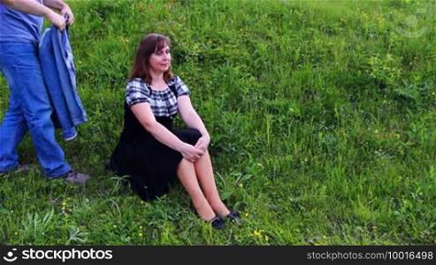 Nice woman sits on grass at hillside, then comes caring man, and sitting alongside