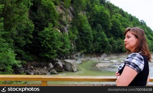 Nice woman stands near railing bridge, then looks at waterfall in Carpathian Mountains