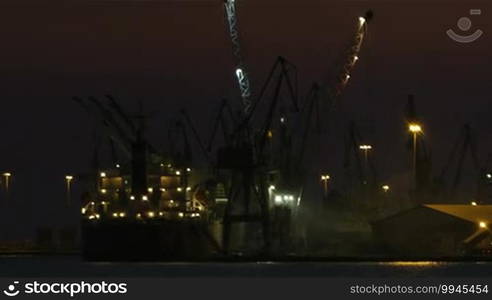 Night view of a harbor in a port with an illuminated cargo ship docked at the wharf being offloaded using cranes