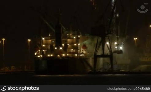 Night view of a harbor in a port with an illuminated cargo ship docked at the wharf being offloaded using cranes