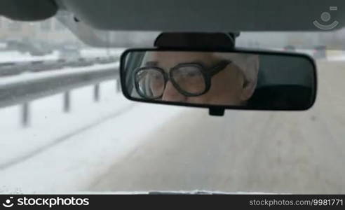 Old man in glasses driving a car. Nasty snowy day and messy roads in the city. Sometimes he looking in the rear mirror