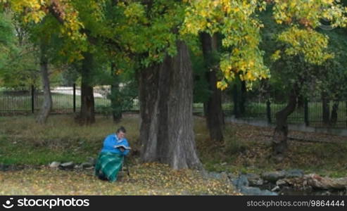 Old woman passing her time by reading a book under an autumn tree in the park. Side view, tilt shot.