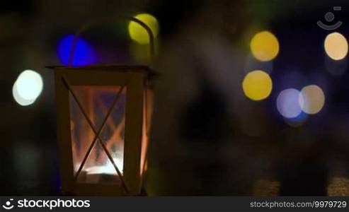 Outdoor lantern with light candle inside swinging in the wind. Blurry colorful lights and people walking in background