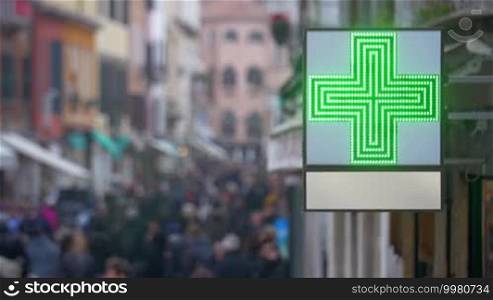 Outdoor pharmacy banner with LED green cross hanging on the building. Defocused crowd of people walking in the street in the background