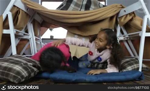 Overexcited two sweet African American little sisters relaxing together at home and tickling each other. Laughing children enjoying leisure in cubby house made of blankets and chairs in domestic interior.