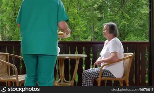 Pan of smiling young nurse serving breakfast to happy senior woman at nursing home. Slow motion