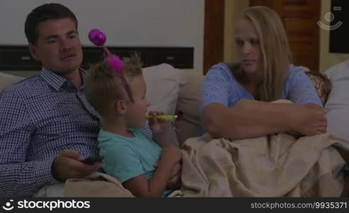 Parents and child at home. Mother and father trying to watch TV while child in funny ears playing with party horn and talking to mom