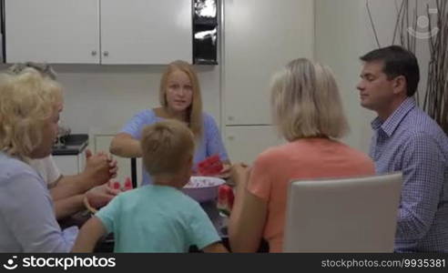 Parents, grandparents, and child eating watermelon in the kitchen. Adults sitting at the table, while the child is running to the mother to get a tasty fruit