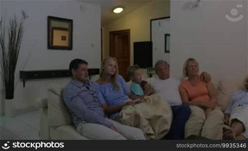 Parents, son, and grandparents sitting on the sofa at home in the evening and watching television. TV program or movie make them laugh