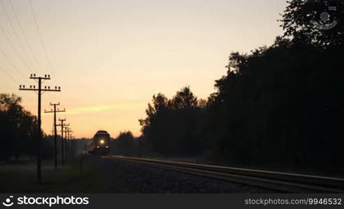 Passenger train with bright headlight passing by slowly in the countryside at sunset.