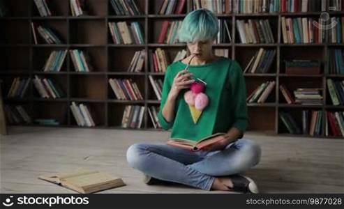 Pensive clever hipster girl in trendy outfit and eyeglasses enjoying reading a novel while sitting on the floor cross-legged at home. Thoughtful young blue-haired woman in spectacles reading a book over bookshelves background. Dolly shot. Slow motion.