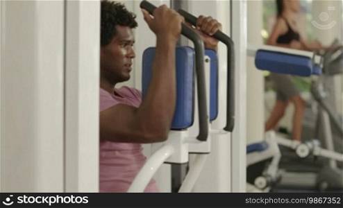 People and sports, young black man exercising pectoral muscles in wellness club, with people working out on cardio bikes in background
