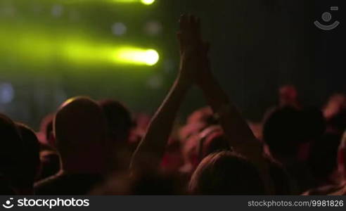 People at the concert. Focus on female hands applauding among the crowd. Digital screen and bright yellow spotlights in the background, then the lights going down