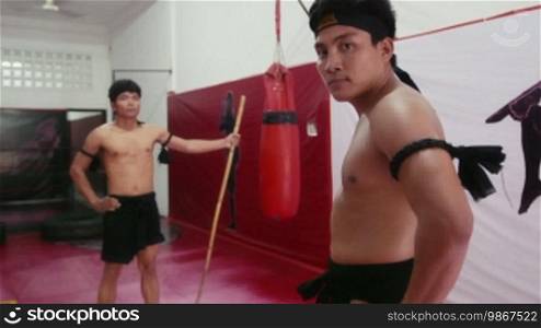 People, athletes, sport activity, workout, combat and extreme sports, Asian men working out for Yuthakun Khom, traditional Khmer martial art of Cambodia. Portrait of trainer looking at camera with student fighting with stick. 17 of 20