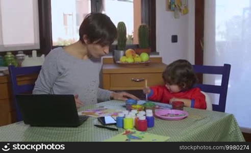 People, family life, fun, lifestyle, childbearing, motherhood. Busy woman talking on the phone, working with PC in kitchen at home, multitasking mother painting with female child, baby girl, daughter