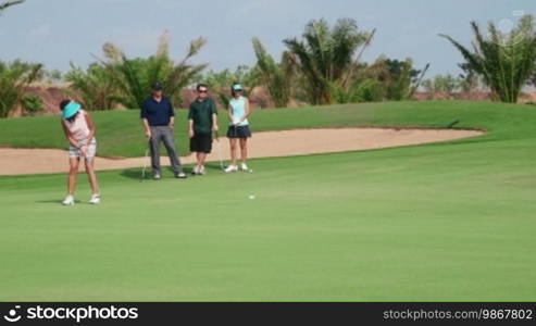 People, sports, leisure activities, recreation and lifestyle, golf in country club during summer holidays. Asian woman hitting ball with iron in golf course near hole, friends watching. 15 of 30