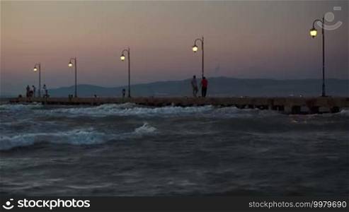 People walking along the pier with lanterns in the evening. Rough sea with strong waves around