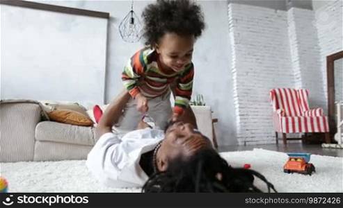 Playful African American dad with dreadlocks lifting his mixed-race little toddler son up while lying on the carpet on the floor in the living room. Happy father playing and having fun with his curly sweet child indoors.