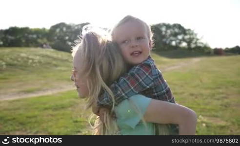 Playful sister piggybacking her toddler brother and running on park lawn. Side view. Positive little girl giving boy piggyback ride while siblings laughing and enjoying leisure while playing together outdoors. Slow motion. Steadicam stabilized shot.