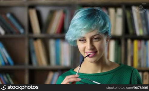 Pondering attractive female journalist with eyeglasses rim in her mouth thinking and generating article ideas for publication and smiling. Thoughtful gorgeous hipster girl pondering preparing a report for homework analyzing information at the library.