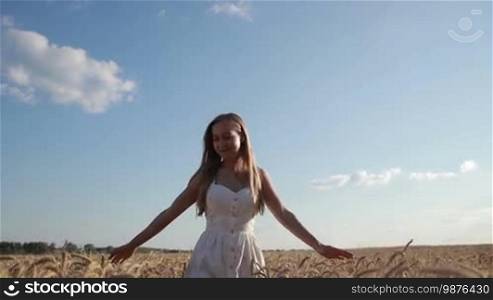 Portrait of a beautiful long blond hair woman in a white summer dress walking through a golden wheat field with a cloudy blue sky in the background. An attractive smiling female with arms outstretched enjoying freedom and happiness in the spikes of the wheat field. Slow motion.