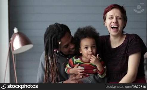 Portrait of a beautiful mixed-race family looking at the camera, smiling, and laughing. Showing positive emotions on their faces. An African American father and a Caucasian mother embracing their adorable mixed-race toddler son at home and smiling. Slow motion.
