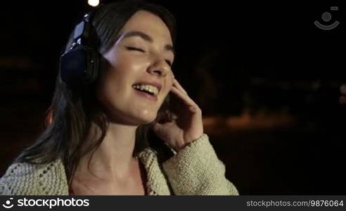 Portrait of a smiling attractive girl with headphones listening to music while spending a great time on a summer night in the city. Gorgeous brunette female in earphones enjoying music at night outdoors. Slow motion. Steadicam stabilized shot.