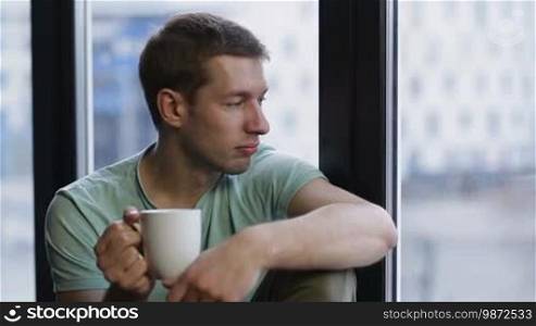 Portrait of a young man sitting by the window with a coffee cup, looking at the city scenery at dawn after waking up early. Handsome guy in casual clothes relaxing in a penthouse after a good night's sleep and watching the sunrise.