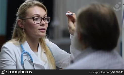 Portrait of an attractive long blonde hair female physician checking pupillary reflex of senior male patient with flashlight in medical office. Attentive female neurologist testing reflexes of elderly man's eyes in clinic during medical checkup.