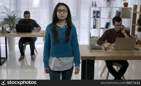 Portrait of attractive Asian female office worker in casual clothes and eyeglasses smiling in freelance office with colleagues working on background. Positive young Asian woman posing at workplace.