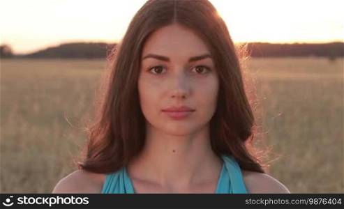 Portrait of charming long hair brunette girl with amazing brown eyes posing in golden wheat field and smiling in glow of setting sun. Sensual young woman looking at camera with soft shy smile over beautiful landscape in summer twilight time.