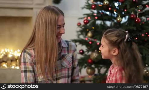 Portrait of cute little girl kissing her beautiful mother at Christmas. Loving daughter giving a kiss to her smiling mom during family Christmas Eve celebration together in decorated living room. Dolly shot.