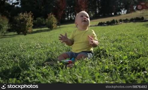 Portrait of cute little infant child sitting on green grassy lawn and clapping his hands with a smile. Sweet toddler baby boy having fun, clapping his hands and enjoying leisure in nature. Slow motion. Steadicam stabilized shot.