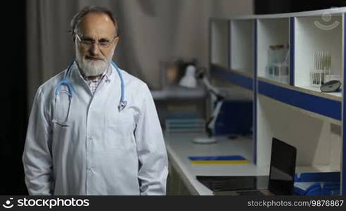 Portrait of friendly senior male doctor with stethoscope in uniform and spectacles standing against medical office background and smiling. Cheerful trusted man medic posing in hospital and looking at cam with radiant smile.