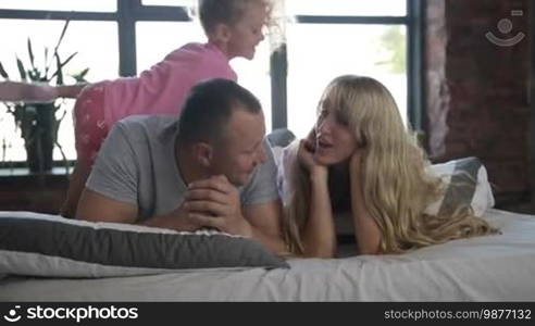 Portrait of happy attractive family with lovely little daughter lying on bed and smiling. Carefree parents with cute girl spending leisure together at home while relaxing at bed in morning. Slow motion. Dolly shot.