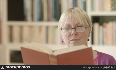 Portrait of happy retired Caucasian woman with eyeglasses reading book at home and smiling at camera