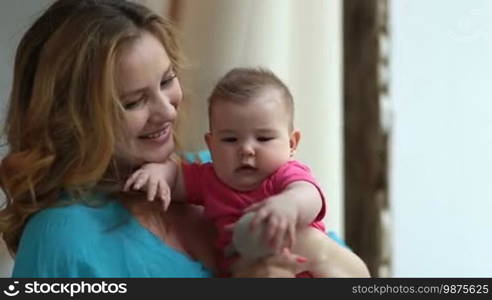 Portrait of happy young mother holding her infant child and playing with toy. Adorable baby girl grasping decorative ball, trying to bite while her caring mom developing palmar grasp reflex of her little child at home.