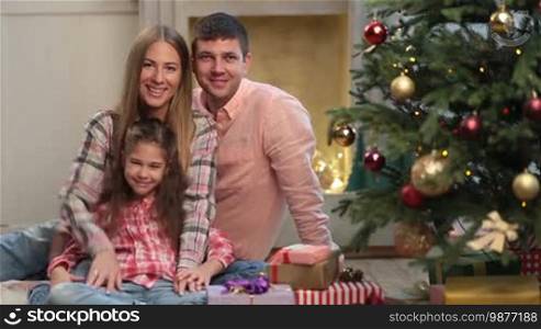 Portrait of positive young family with lovely daughter waving hands and smiling over Christmas decorated room background. Attractive parents and little girl sitting together near Christmas tree during winter holidays, looking at camera and waving hands.