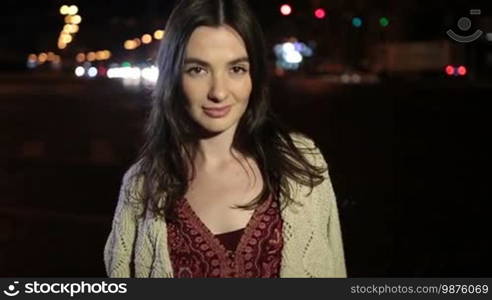 Portrait of seductive brunette woman flirting with camera and looking at someone temptingly over night city streetlights and traffic background. Sexy beautiful girl with enigmatic look posing on street at night. Slow motion. Stabilized shot.