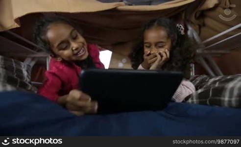 Portrait of smart African American kids using a digital tablet PC in a domestic interior. Adorable mixed-race little girls playing online games on a touchpad while lying in a cubby house made of blanket and chairs. Close-up. Low view.
