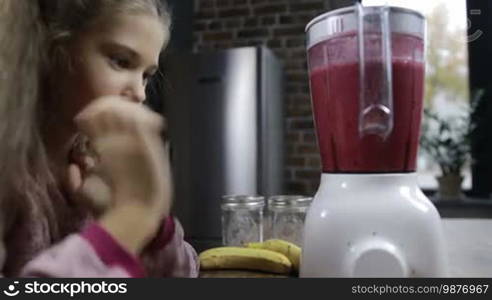 Portrait of smiling little girl preparing fresh berry smoothie in blender at kitchen. Adorable girl with long brown hair watching the process of blending healthy berry smoothie in blender shaker jug and smiling. Dolly shot.