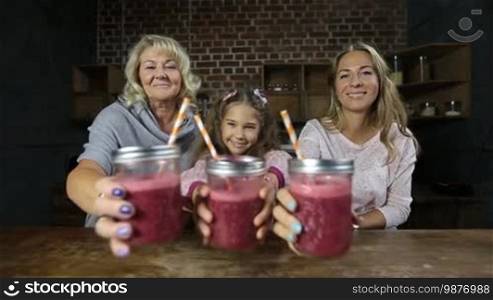 Portrait of three generation family holding mason jars of fresh red berry smoothies with drinking straws and smiling. Beautiful happy women offering glass jars of detox weight loss drinks over modern domestic kitchen background.